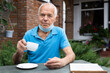 © caftor - Elderly man in protective mask with cup of coffee or tea in his hand sits at table of street cafe and reads menu
