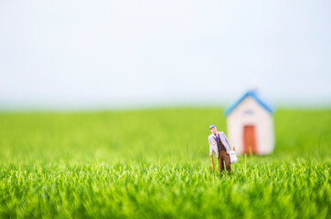 Miniature elderly man carry grocery bag walking in the field with space on blurred house background, selective focus, retired concept