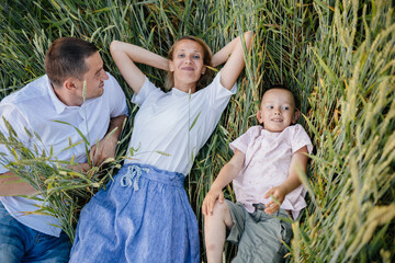 Above view of happy young family with little son lying and resting among green wheat field at summer evening at countryside together..