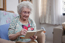 Elderly Lady At Home Sitting In A Chair Reading A Book Using A Magnifying Glass