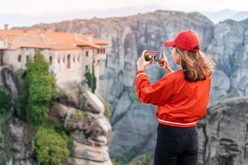 Wall Mural - Tourist girl taking photos on smartphone of the scenic Meteora Monstery of Varlaam near Kalabaka town. Sightseeing and travel blogger concept
