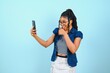 Happy cheerful Afro American woman looks at screen of smart phone enjoys online chatting types text message surfs social networks dressed casually poses against blue background. Technology concept