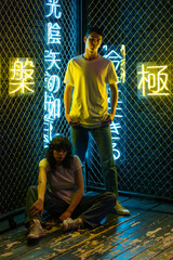 Wall Mural - Young couple in white t-shirts stand against the background of a metal grid with neon symbols.