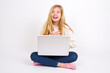 © Jihan - caucasian teen girl sitting with laptop in lotus position on white background keeps hands crossed, laughs out loud at good joke, wears casual clothes.