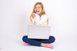 © Jihan - Dreamy caucasian teen girl sitting with laptop in lotus position on white background with pleasant expression, closes eyes, keeps hands crossed near face, thinks about something pleasant