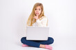 © Jihan - caucasian teen girl sitting with laptop in lotus position on white background makes hush gesture, asks be quiet. Don't tell my secret or not speak too loud, please!