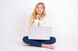 © Jihan - Dreamy charming caucasian teen girl sitting with laptop in lotus position on white background with pleasant expression, keeps hands crossed near face, excited about something pleasant.
