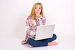 © Jihan - Oh! I think I said it! Close up portrait caucasian teen girl sitting with laptop in lotus position on white background cover open mouth by hand palm, look at camera with big eyes.