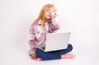 © Jihan - Frustrated caucasian teen girl sitting with laptop in lotus position on white background holding hand on forehead being depressed regretting what he did having headache, looking stressful.