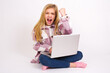 © Jihan - Fierce caucasian teen girl sitting with laptop in lotus position on white background holding fist in front as if is ready for fight or challenge, screaming and having aggressive expression on face.