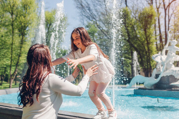 Young mother and her daughter having fun by fountain in spring park. Girl jumps in mom's hands. Mothers day