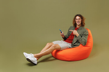 Full size body length young brunet curly man 20s wear khaki shirt sit in bag chair hold in hand use pointing index finger on mobile cell phone isolated on plain olive green background studio portrait