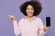 © ViDi Studio - Young woman of African American ethnicity in pink striped shirt hold in hand use mobile cell phone with blank screen workspace area point finger aside isolated on plain pastel light purple background.