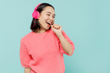 Young Smiling Happy Woman Of Asian Ethnicity 20s Wearing Pink Sweater Headphones Listen To Music Sing Song In Microphone Isolated On Pastel Plain Light Blue Color Background. People Lifestyle Concept.