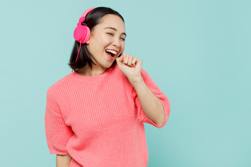 Wall Mural - Young smiling happy woman of Asian ethnicity 20s wearing pink sweater headphones listen to music sing song in microphone isolated on pastel plain light blue color background. People lifestyle concept.