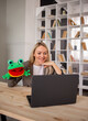 Portrait of a female speech therapist in a jacket remotely conducts a lesson with a laptop. A female speech therapist shows an exercise with a knitted frog toy
