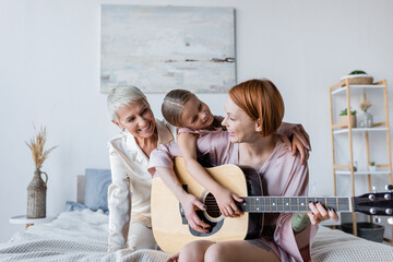 Positive woman playing acoustic guitar near adopted daughter and girlfriend on bed