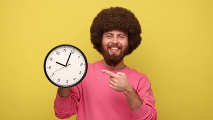 Wall Mural - Bearded hipster man with Afro hairstyle holding pointing at big wall clock, looking at camera with pleasant smile, time to go, wears pink sweatshirt. Indoor studio shot isolated on yellow background.