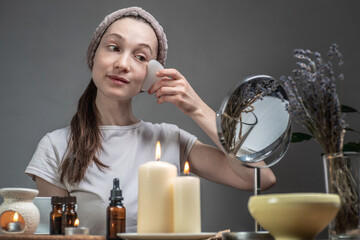 Wall Mural - Woman is looking in the mirror and doing a massage with a gouache scraper. Candles and an aroma lamp with essential oils on the table. Concept of facial care, aromatherapy, morning and evening rituals