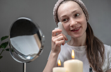 Wall Mural - Woman is looking in the mirror and doing a massage with a gouache scraper. Candles and an aroma lamp with essential oils on the table. Concept of facial care, aromatherapy, morning and evening rituals