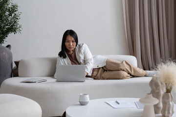 Aufkleber - Serious young Asian female manager looking at laptop screen while lying on white sofa in living-room and analyzing online data