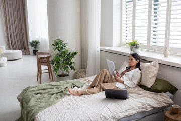 Aufkleber - Young confident Asian female accountant in casual clothes sitting on bed and looking through financial papers while working from home