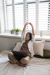 Aufkleber - Young tired employee with laptop on crossed legs stretching raised arms while sitting on double bed and working online from home office