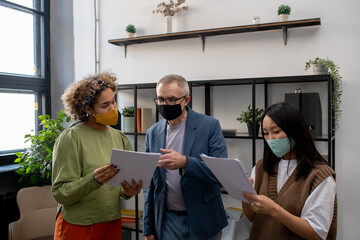Aufkleber - Mature businessman in mask pointing at document during discussion with young biracial female colleague at working meeting in office