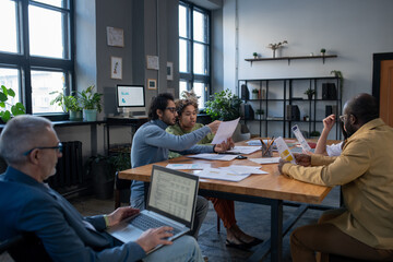 Aufkleber - Group of intercultural economists discussing financial papers by large table while mature businessman with laptop preparing presentation
