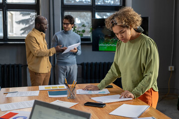 Aufkleber - Young female economist looking through financial data and analyzing it by table against two male colleagues preparing report