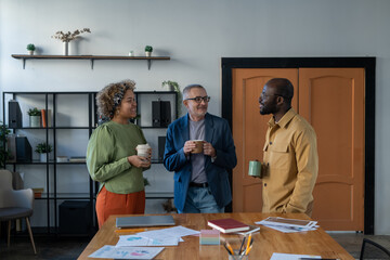 Aufkleber - Three confident intercultural colleagues having drinks and talking at break while standing by large table with financial documents in office