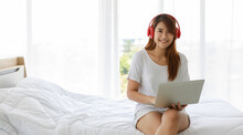 Portrait Shot Of Asian Young Beautiful Sexy Sensual Long Brown Hair Female Model In Casual Outfit Smiling Sitting On Bed Wears Headphone Listen To Music Song Playlist From Laptop Computer In Bedroom