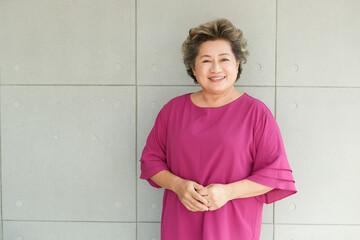 Portrait shot of Asian old senior chubby female retirement pensioner gray short hair grandmother model in casual pink outfit standing smiling look at camera holding hands together on wall background