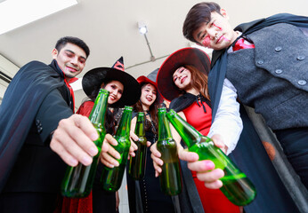 Wall Mural - Portrait shot group of Asian young happy male and female friends in Halloween witch sorcerer ghost and skeleton costume with tall hat sitting on sofa holding beer bottles cheers together in party