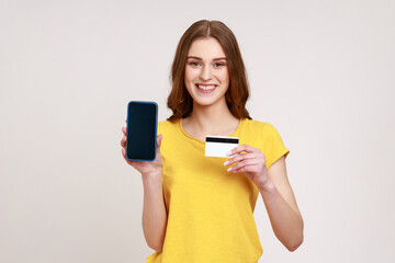 Wall Mural - Cheerful teenager girl in yellow casual style t-shirt holding and showing empty display smartphone and credit card, satisfied with easy mobile banking. Indoor studio shot isolated on gray background.