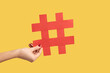 Closeup of woman hand holding large red paper hashtag symbol, hash sign of famous media content, social media marketing and blog promotion. Indoor studio shot isolated on yellow background.
