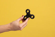Closeup Side View Profile Portrait Of Woman Hand Holding Black Fidget Spinner, Stress Relieving Toy. Indoor Studio Shot Isolated On Yellow Background.
