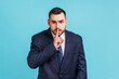 © khosrork - Portrait of bearded businessman wearing official style suit holding finger near lips and showing silence sign, keeping secrets, deceit. Indoor studio shot isolated on blue background.