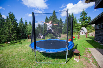 Wall Mural - Happy little girl jump on trampoline in backyard. Child fun outdoor. Summer day. Blue sky. Nature landscape in background.