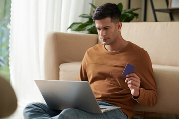 Young man sitting on the floor in the room with laptop on his knees paying purchases online using his credit card