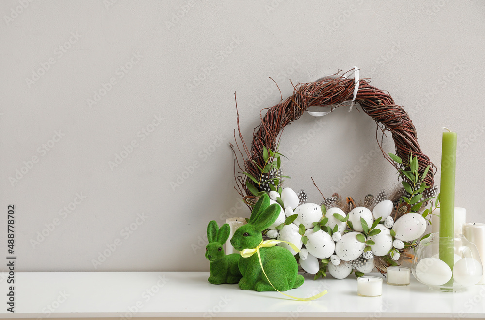 Beautiful Easter wreath, toys and candles on table near light wall