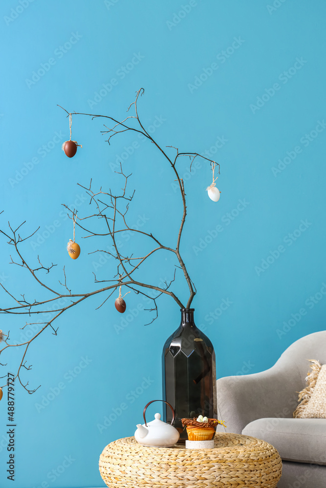 Vase with tree branches decorated for Easter celebration, delicious bun and teapot on table near blue wall