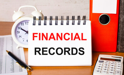 Fototapete - On the table are reports, a white alarm clock, a calculator, folders for papers, a pen and a white notebook with the FINANCIAL RECORDS. Business concept