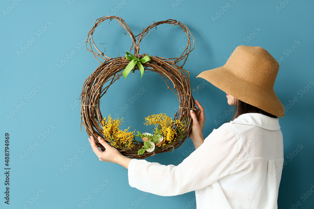 Woman with stylish hat hanging beautiful Easter wreath on color wall