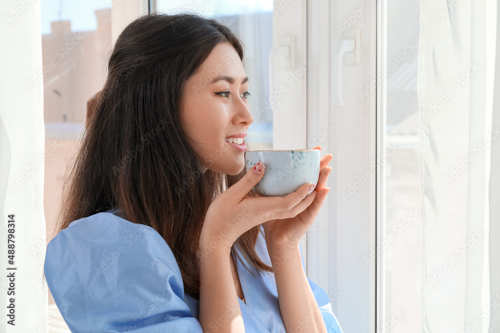 Pretty young Asian woman drinking coffee near window
