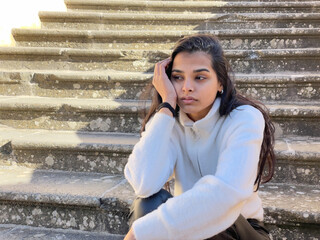 Sad looking young woman sitting outside and holding her head while looking away