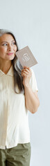 Wall Mural - Thoughtful hoary haired Asian lady customer holds gift card standing on light grey background in studio. Shopping certificate