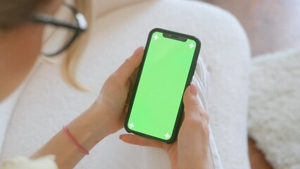 Young woman sitting on sofa at home and uses smartphone with green screen Mock-up. 4k