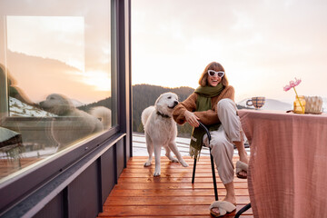 Woman playing with her dog while resting on terrace of tiny house in the mountains, enjoying beautiful landscape during sunrise. Concept of small modern cabins for rest and escape to nature