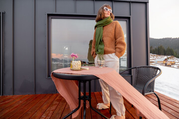 Wall Mural - Young stylish woman standing on terrace of tiny house in the mountains, enjoying beautiful landscape during sunrise. Concept of small modern cabins for rest and escape to nature